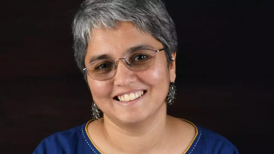 Janaki Srinivasan has short grey hair and glasses. She is wearing a navy blue dress with embroidered details around the neckline, and large silver earrings.