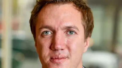 Neil has brown hair, green eyes and is wearing a pale blue shirt under a brown woollen jacket.  