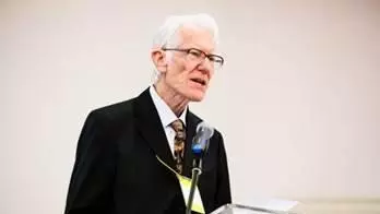 Arthur Stockwin has grey hair and wearing glasses. He's standing at a podium speaking into a microphone. He's wearing a white shirt, tie and smart black jacket. 