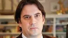 dimitar bechev has black hair and is looking at the camera. He's dressed in a white shirt, deep red tie, and navy blue jumper under a dark brown jacket. 