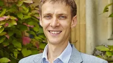 Justin has short light brown hair and is smiling at the camera. He's wearing a shirt, with vertical blue and white stripes, under a grey jacket. 