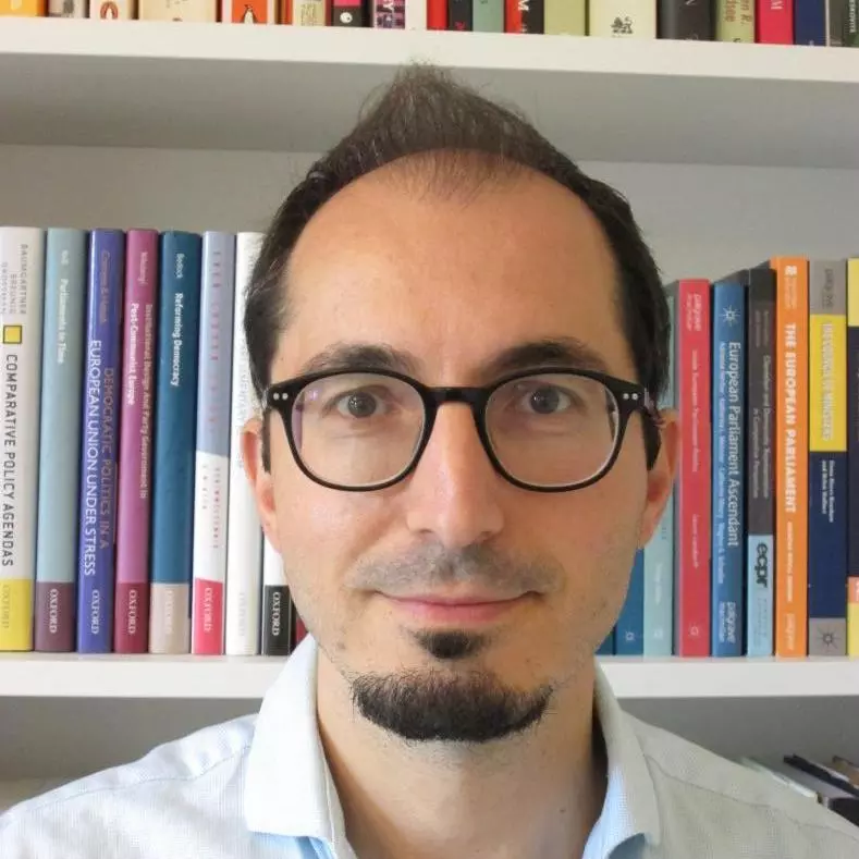 Dr Chiru has brown hair and a short beard. He is wearing dark round glasses and standing in front of a bookshelf