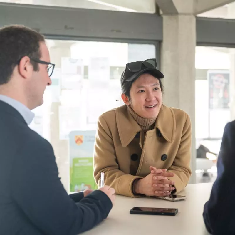 Two students and a teacher stand around a table, chatting. One student is facing the camera, wearing a tan jacket and black cap. 