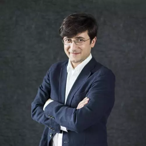Dr Maziashvili has short dark hair. He is wearing a navy blue suit with a white shirt, standing with his arms folded and smiling