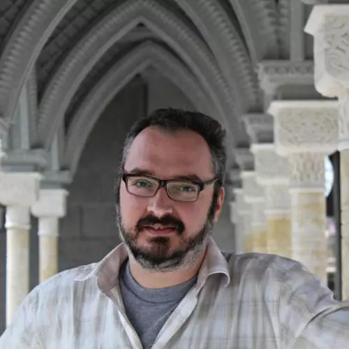David has short dark hair, short beard and glasses. He is standing beneath a row of ornate stone arches