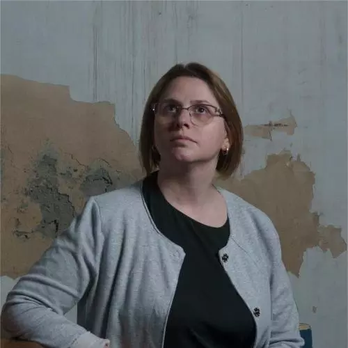 Tea Kamushadze has short brown hair and glasses. She is wearing a grey cardigan and black dress and standing in front of a distressed wall