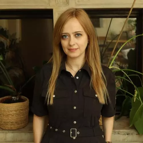 Esra Akgemci has long blonde hair. She is wearing a black jumpsuit and standing in front of some plants