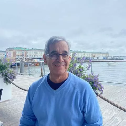 Jesus Velasco has short grey hair and glasses. He is wearing a light blue jumper and sat at a waterfront