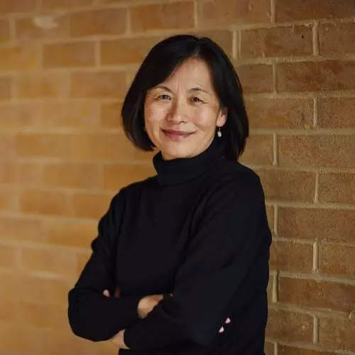 Professor Sako has dark bobbed hair and is wearing a black top. She is leaning against and wall with her arms folded, smiling