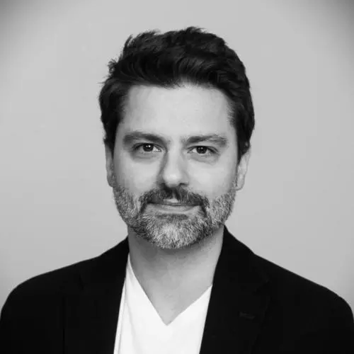 A black and white photo of Felipe who has short dark hair and grey beard. He's wearing a white v neck t shirt under a smart black jacket. 