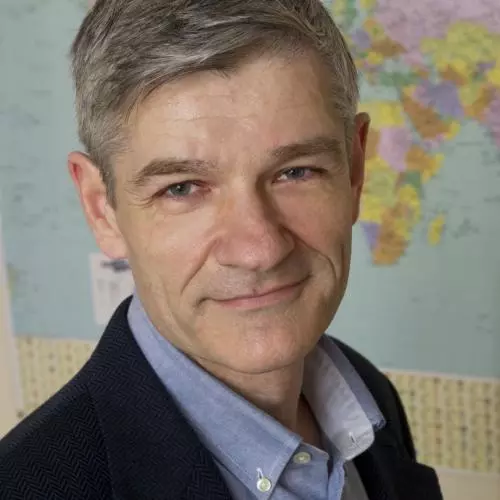 Roy Allison has short grey hair and is looking towards the camera. He's wearing a pale blue shirt with smart navy blue jacket. There is a world map behind him. 