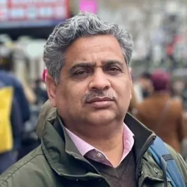 Professor Raja has grey curly hair and a moustache. He is wearing a pink shirt under a grey jumper, and a dark green coat.  
