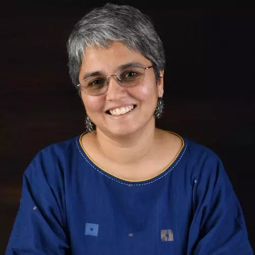 Janaki Srinivasan has short grey hair and glasses. She is wearing a navy blue dress with embroidered details around the neckline, and large silver earrings.
