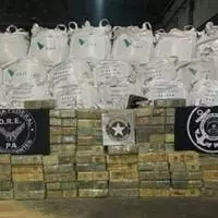 A wall of white industrial bulk bags stacked in rows behind neatly piled brown wrapped packages. Two armed officers are standing facing the packages.