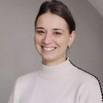 marija has dark hair tied back behind her head. She's smiling at the camera and wearing a white jumper