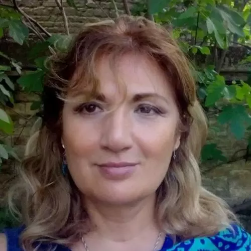 leila alieva has shoulder length wavey blonde hair and is wearing a blue patterned top. She's standing in front of a brick wall and shrubs. 