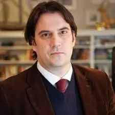 dimitar bechev has black hair and is looking at the camera. He's dressed in a white shirt, deep red tie, and navy blue jumper under a dark brown jacket. 