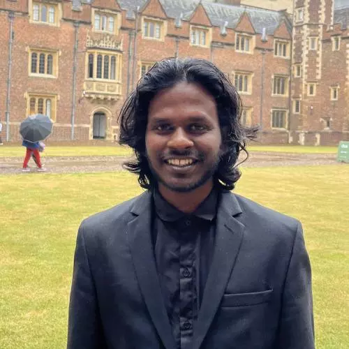 Roya has black wavey hair and a bead. He's dressed in a dark grey shirt and jacket as is standing in a college quad.