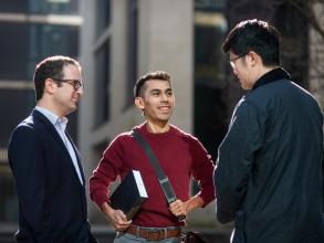 Two students and a teacher in a group, standing and chatting outside department buildings. 