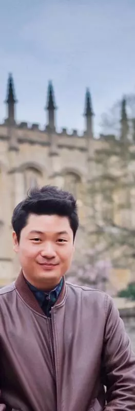 A man with dark hair and wearing a brown jacket over a blue shirt is standing in an Oxford college garden with a church behind him. 