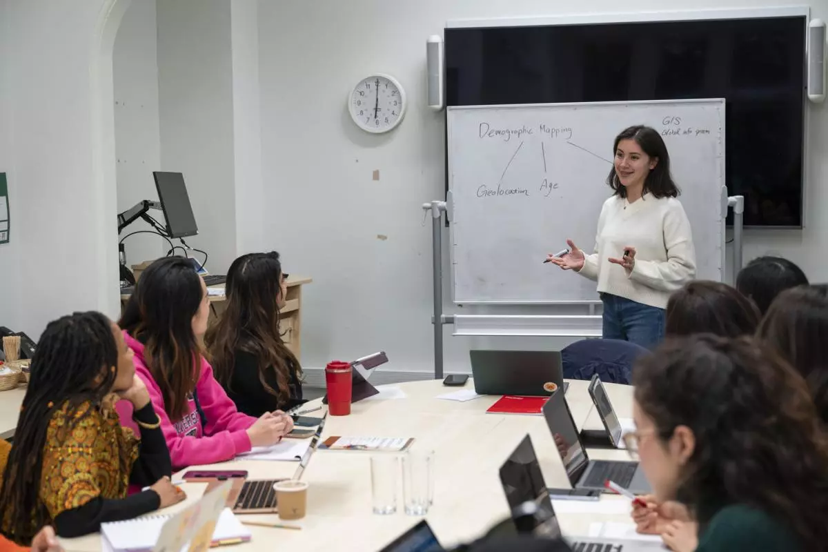 A group of people sitting around a large table, looking at a women in a white jumper standing in front of a whiteboard.