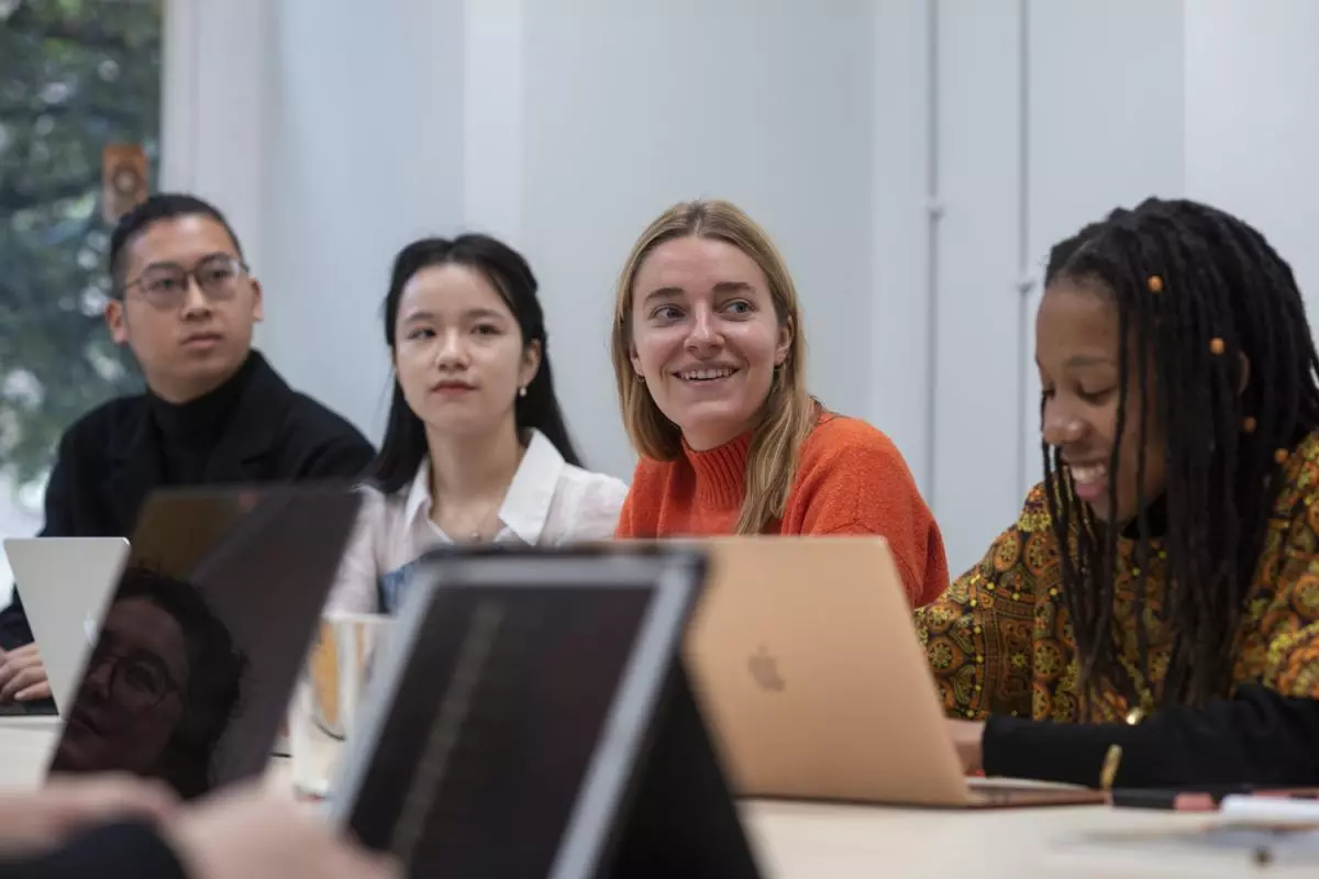 Four people sitting on one side of a meeting table, all at laptops. Three are looking to the right and smiling. In the foreground on the right, another smiles and looks at her notes.