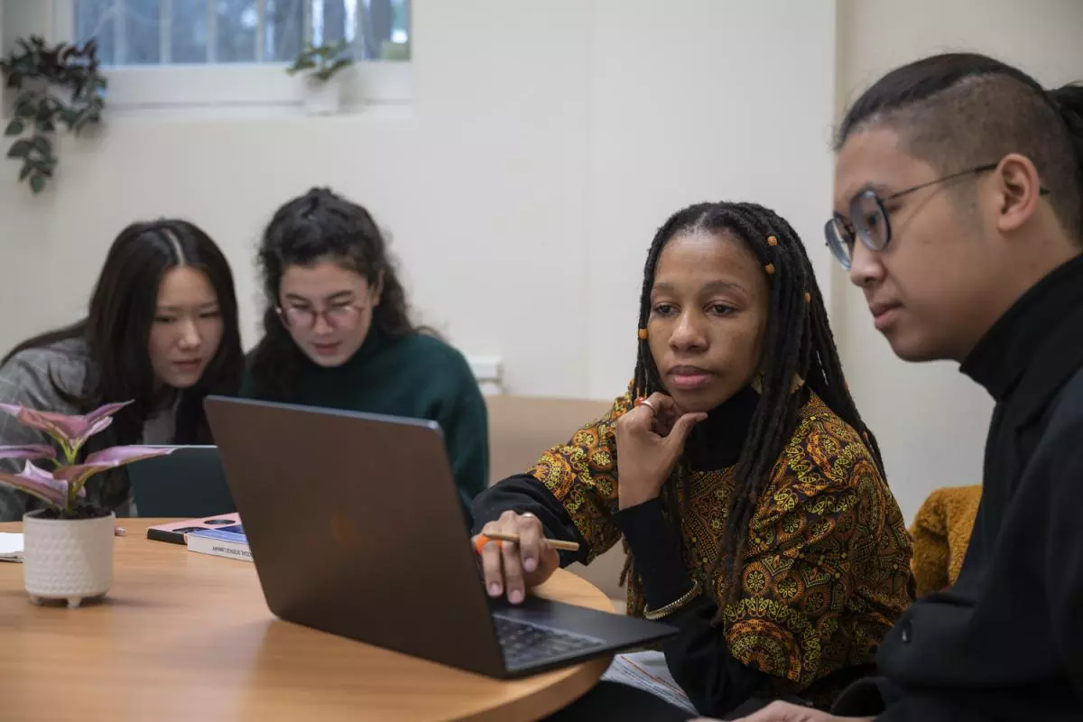 Two students sitting at a table, looking at a laptop screen together. Two other students are sat at another laptop in the background. 
