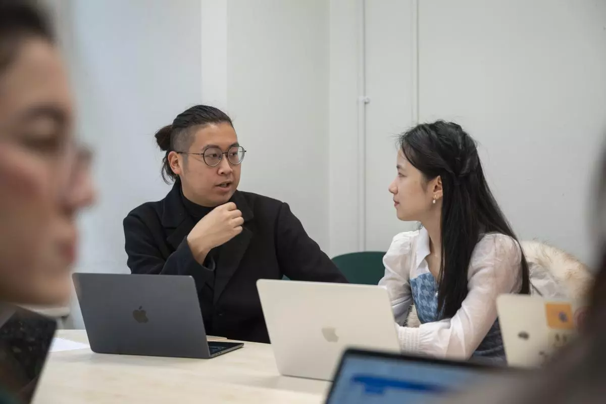 Two students sat a a desk with laptops, turned towards each other and chatting