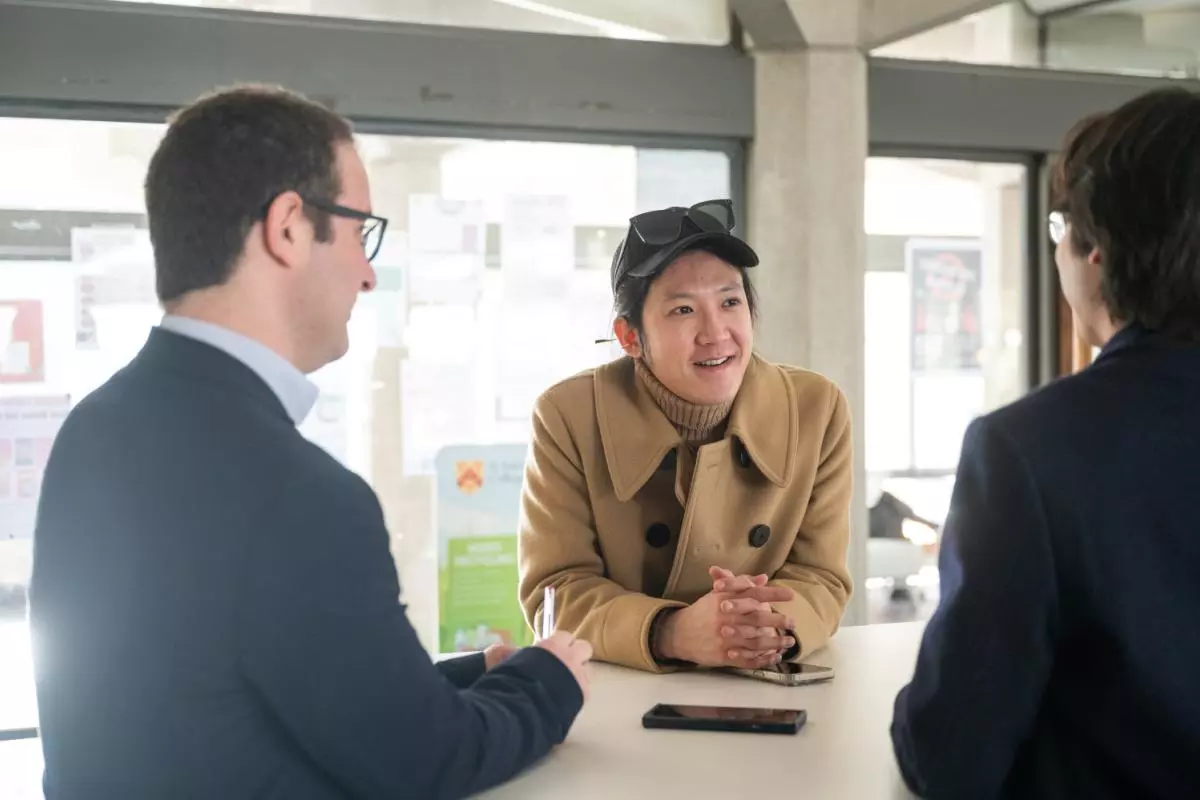 Two students and a teacher stand around a table, chatting. One student is facing the camera, wearing a tan jacket and black cap. 